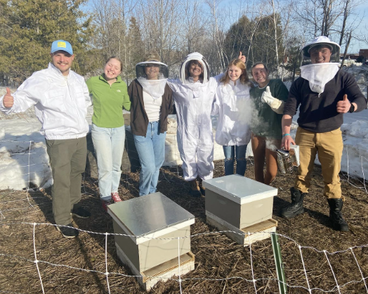 7 members of Helping Hives wearing bee keeping suits posing with two bee hives. 
